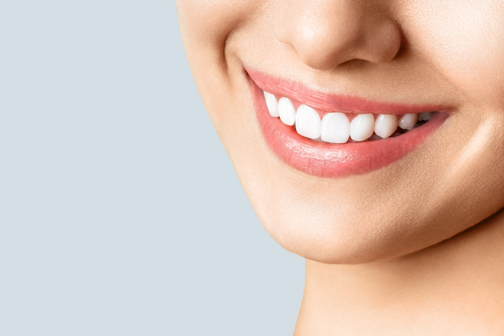 Close-up of a smiling persons mouth showing straight, white teeth and smooth skin, with a light background.