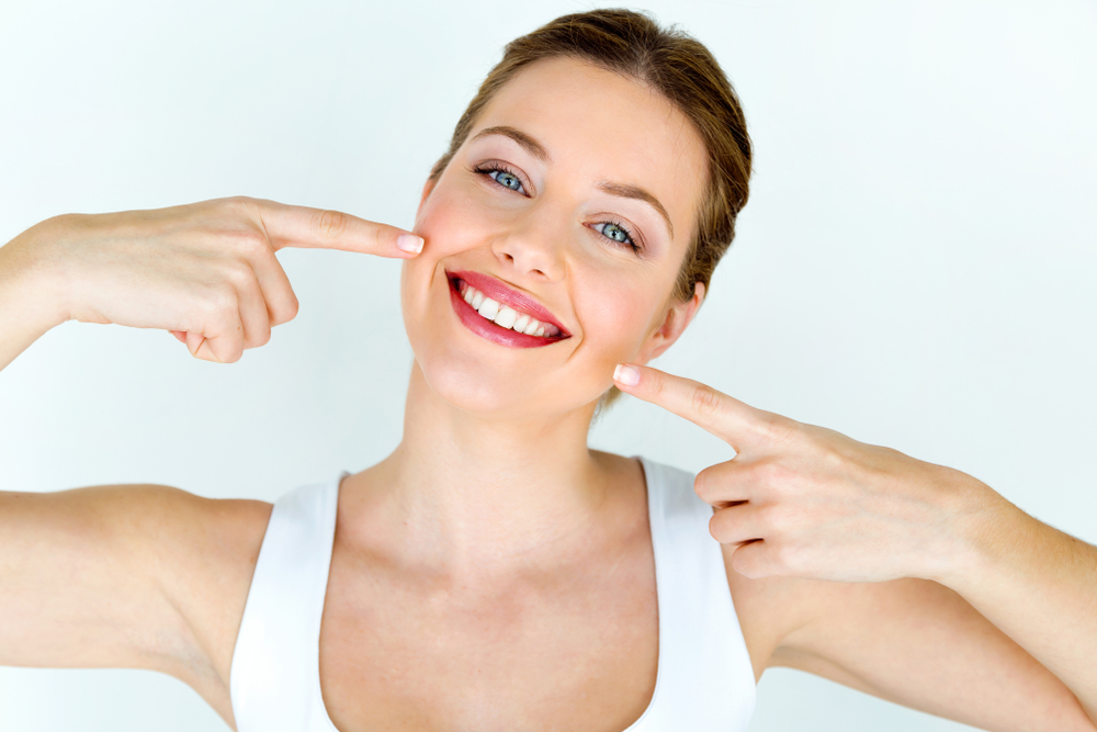 Smiling woman with light skin and blue eyes points to her teeth with both index fingers. She is wearing a white top and standing against a plain, light background.